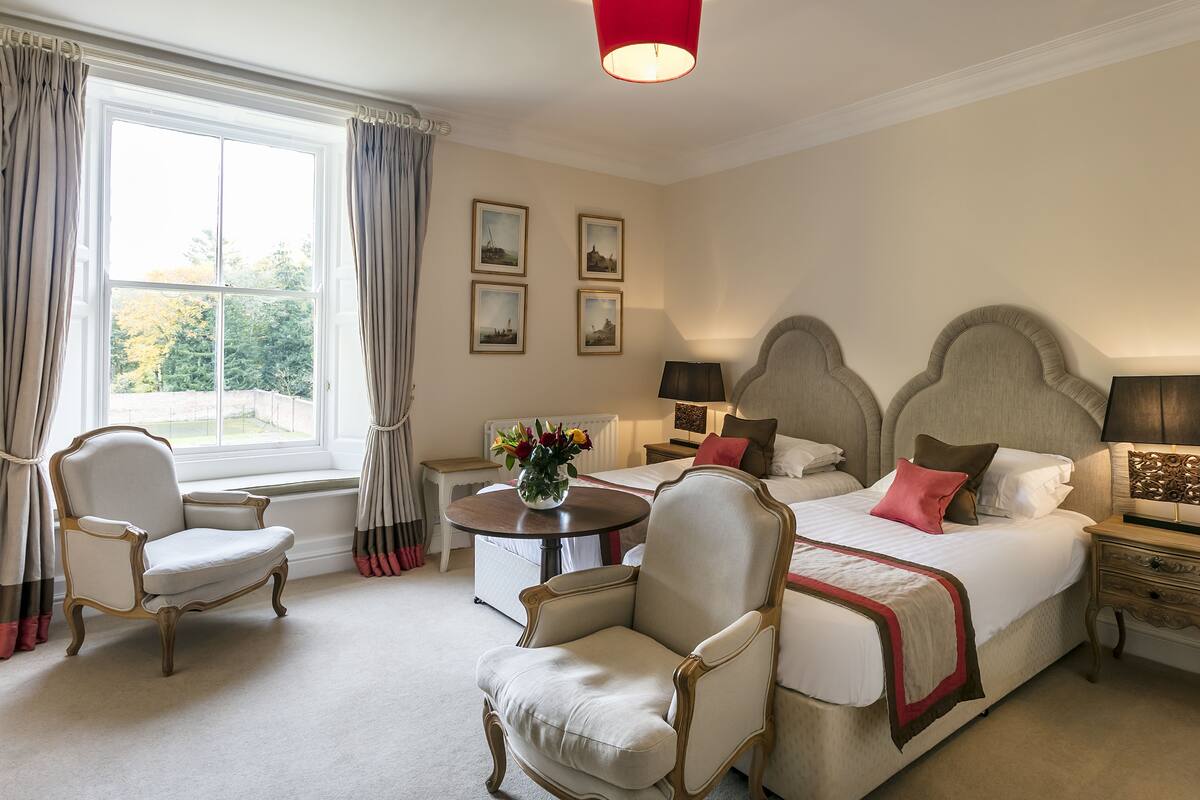 A hotel room with a large bed, two armchairs, a small table with a vase of flowers, and a window with curtains.