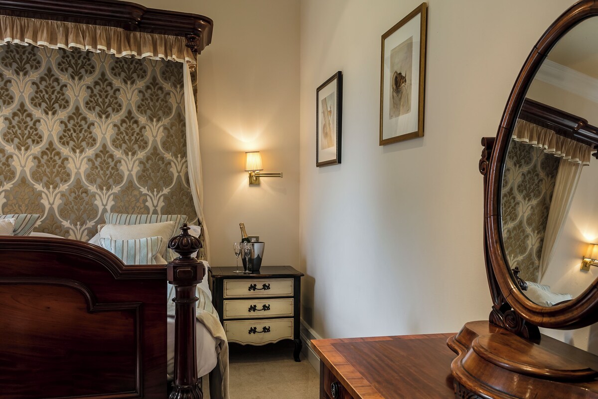 A bedroom with a four-poster bed, a bedside table with drawers, a mirror, and framed pictures on the wall.