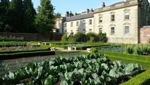 A large stone building with multiple windows and a symmetrical garden with neatly trimmed hedges and a central fountain. - Eshott Hall (Morpeth)