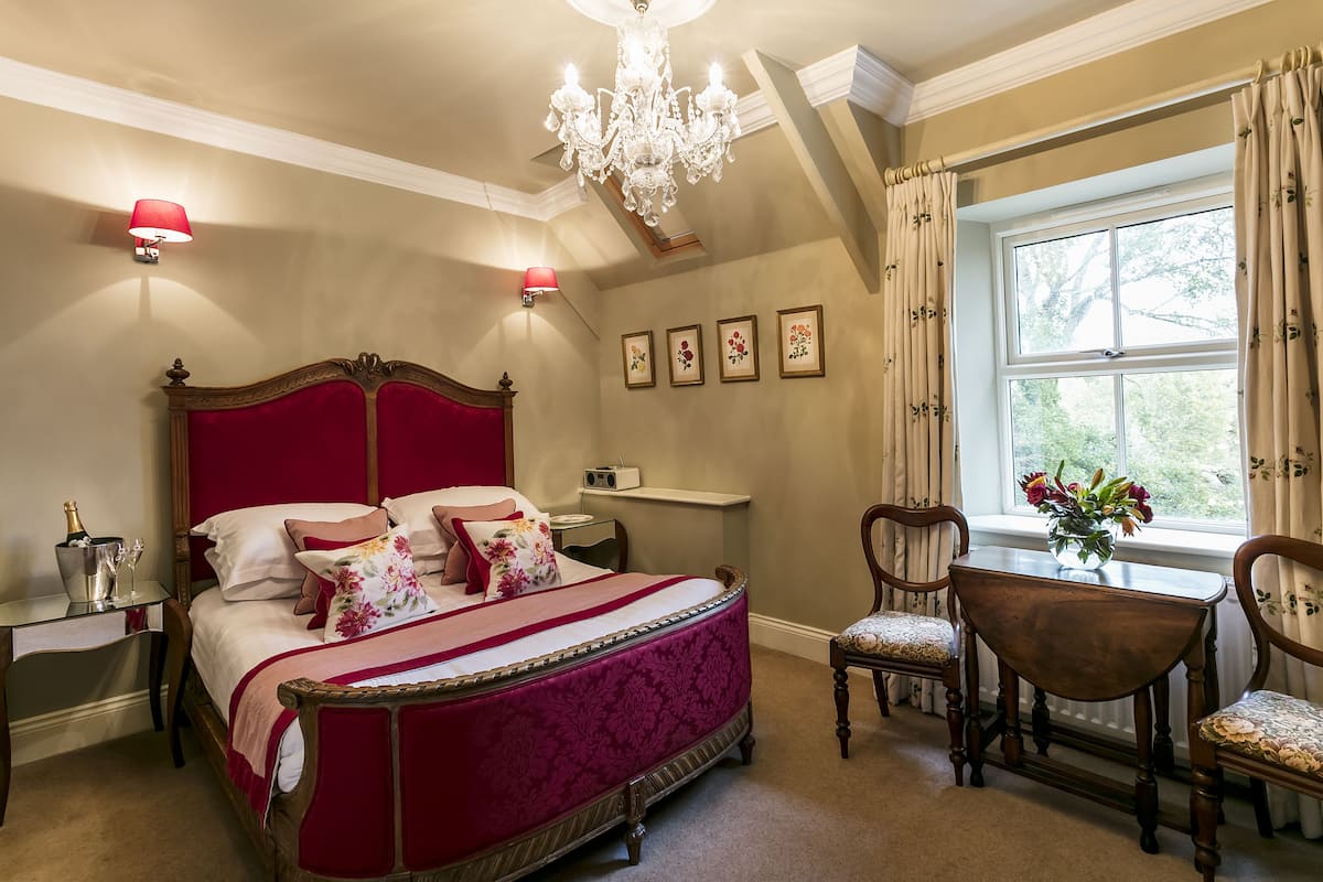 A bedroom with a large bed, a chandelier, a desk with a lamp, and a window with curtains.