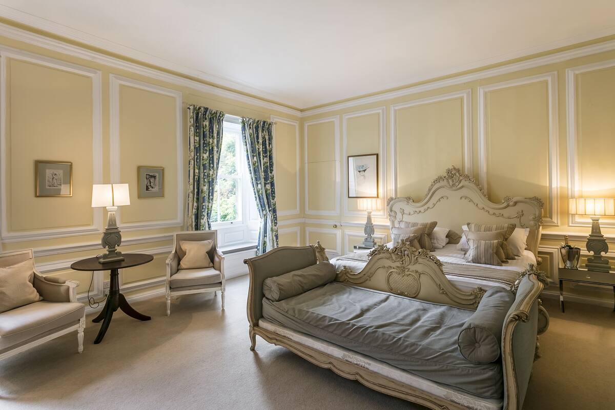 A classic bedroom with a large bed, two bedside tables with lamps, a chair, and a window with curtains.