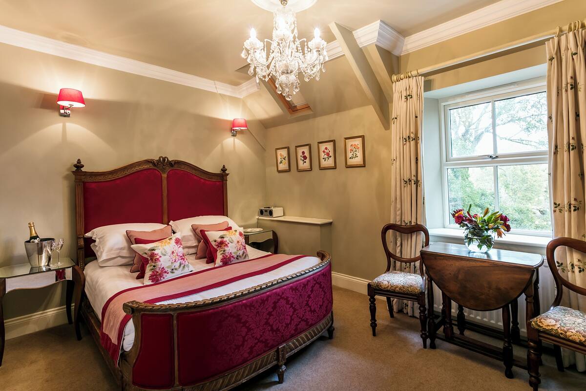 Quarto com uma cama grande, um candelabro, uma escrivaninha com luminária e uma janela com cortinas.