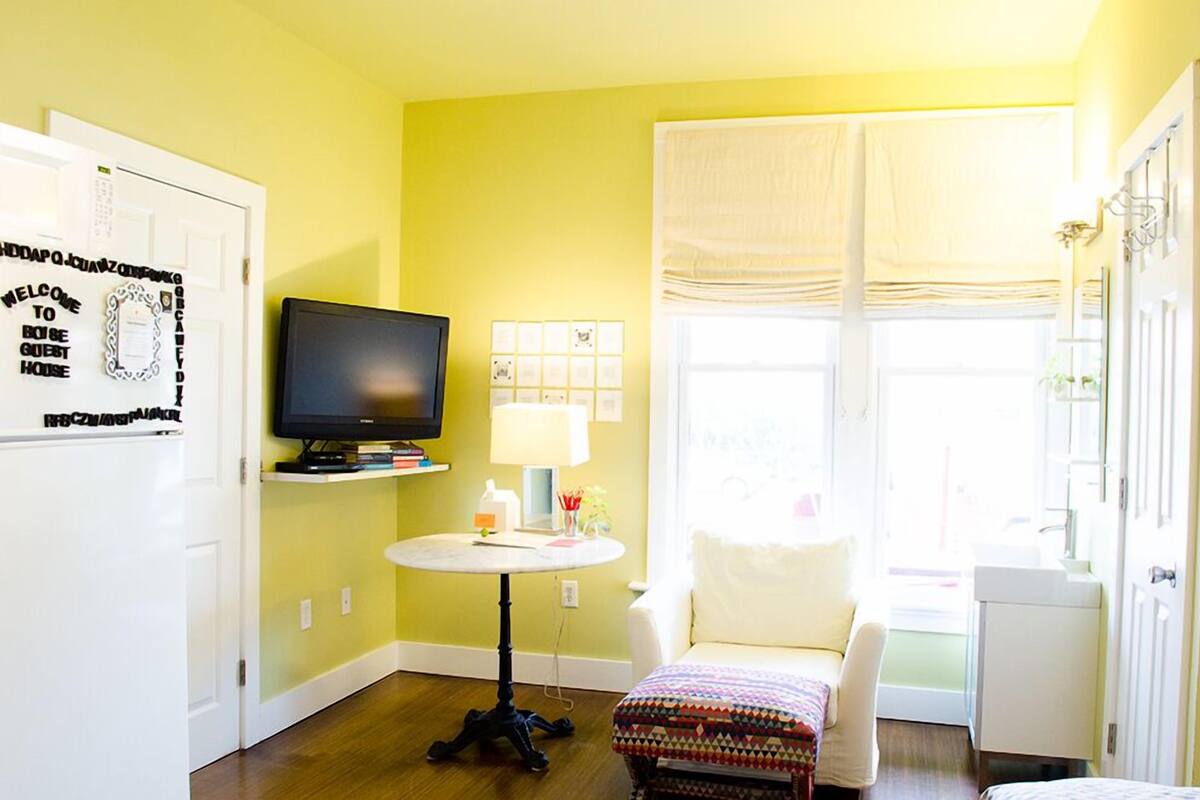 A room with a television on a stand, a small round table with a lamp, a chair, and a window with curtains.