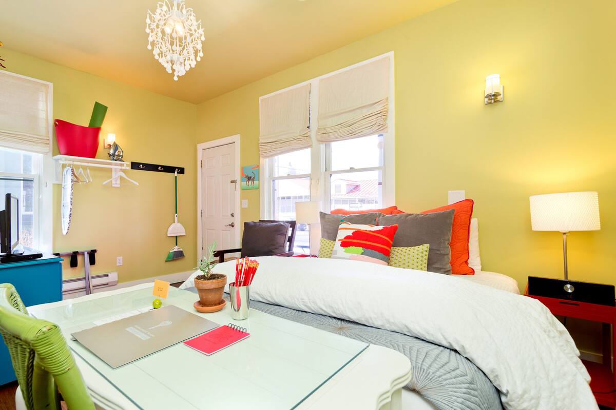 A hotel room with a bed, a desk with a laptop, a chair, a potted plant, and a window with curtains.