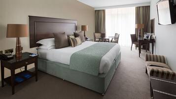 A hotel room with a large bed, two bedside tables with lamps, a desk with a chair, a TV mounted on the wall, and a window with curtains.
