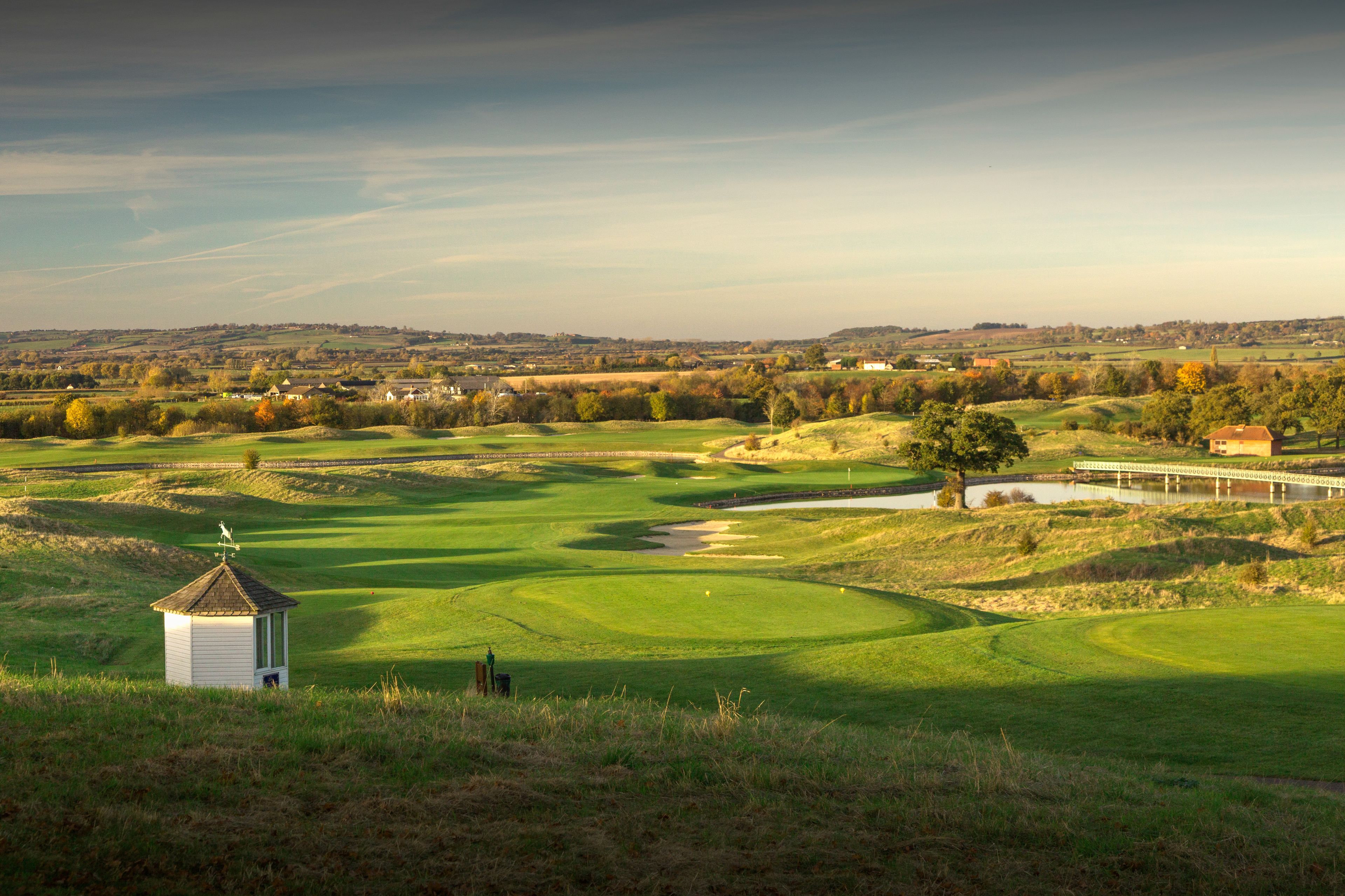 A golf course with a small white building, a bridge, and a distant view of houses and trees.