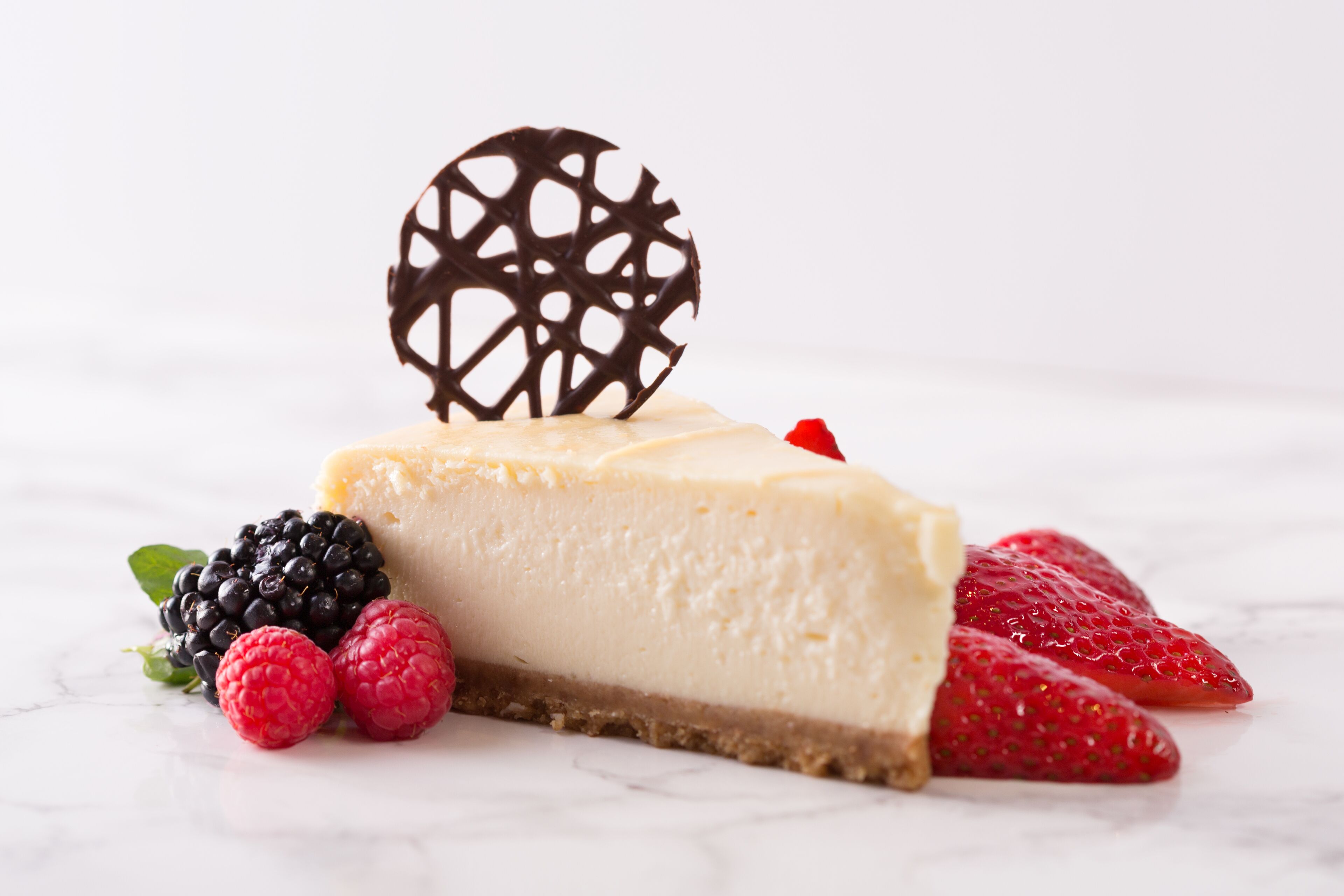 A slice of cheesecake with a decorative chocolate piece on top, accompanied by strawberries and raspberries.