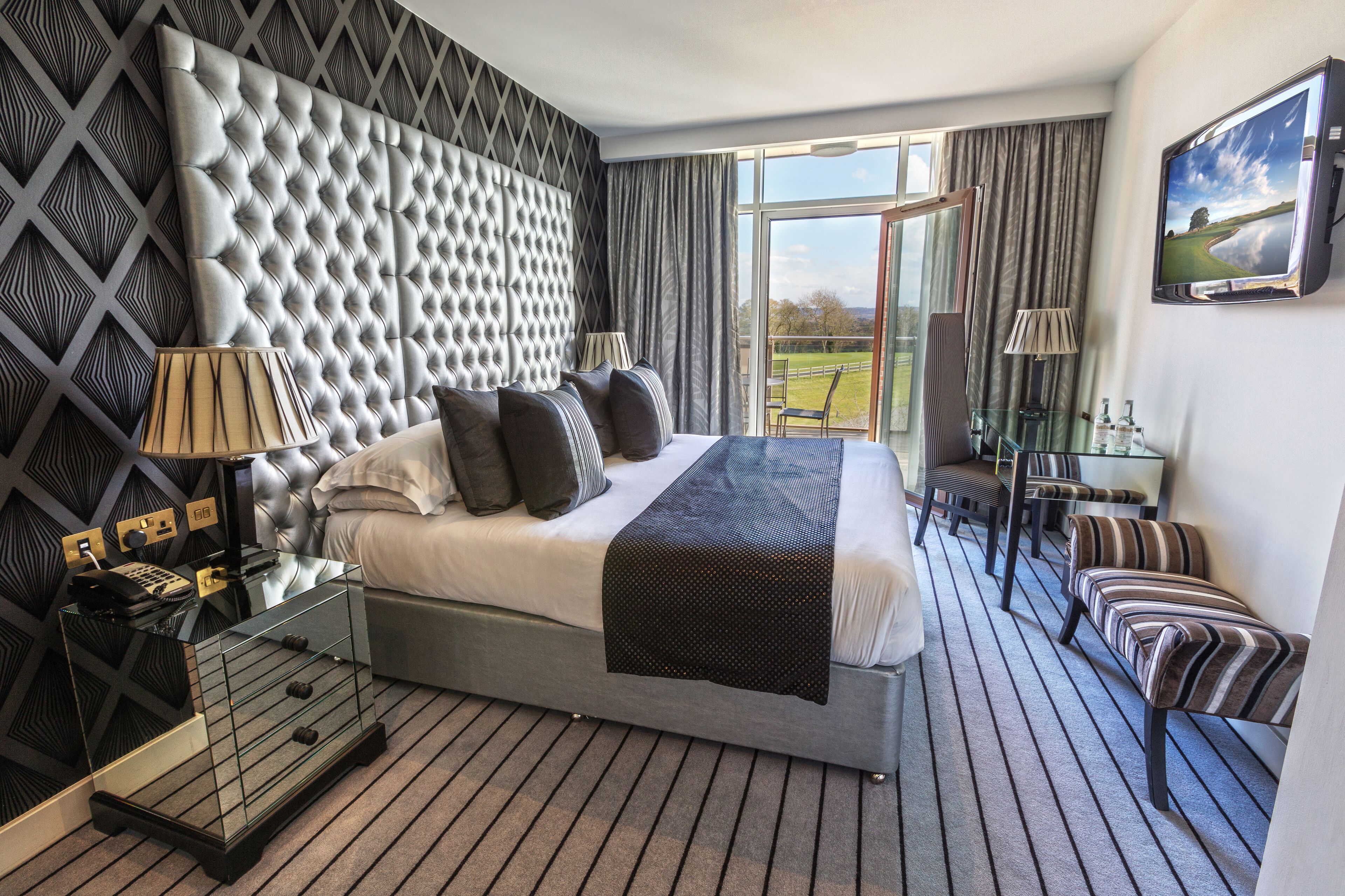 A modern hotel room with a large bed, a glass coffee table, a seating area with chairs, and a view of the outdoors.