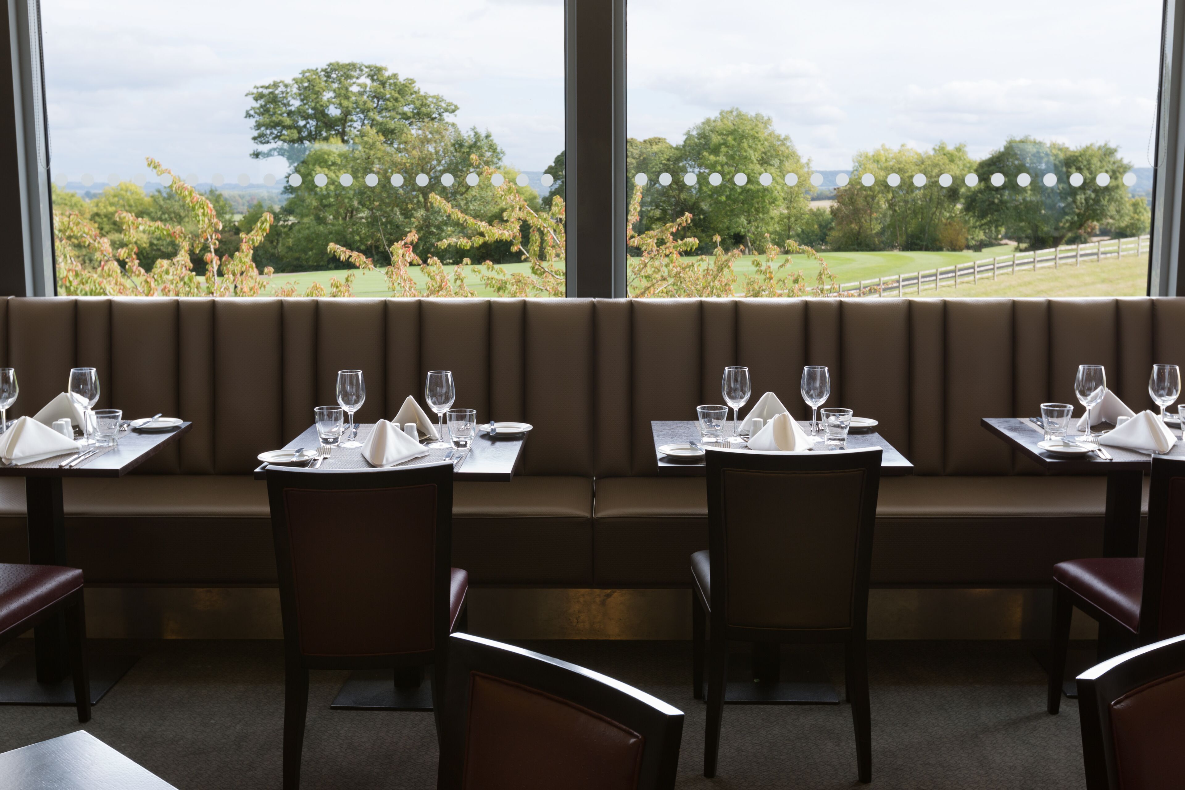 A restaurant with a long bench seating area, tables with chairs, and a view of a green landscape through large windows.