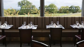 A restaurant with a long bench seating area, tables with chairs, and a view of a green landscape through large windows.