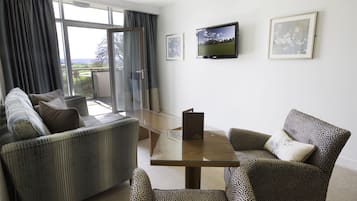 Un salon comprenant un canapé, des fauteuils, une table basse avec un plateau en verre et un téléviseur fixé au mur.