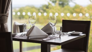 Una mesa de comedor dispuesta con una servilleta blanca, dos copas de vino, un vaso de agua, cubiertos y un plato, con vistas a la vegetación y luces en el fondo.