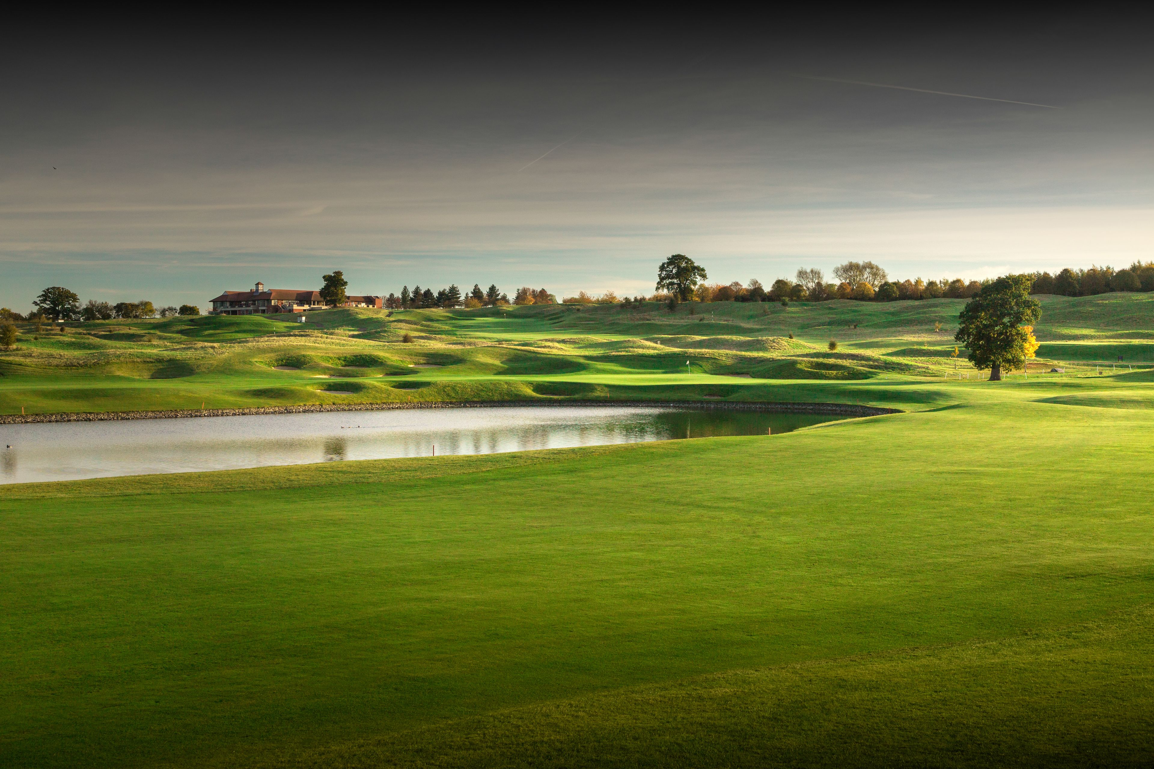 Een golfbaan met glooiende groene heuvels, een vijver en een gebouw op de achtergrond.