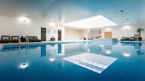 Una piscina interior con agua cristalina azul, un techo iluminado por el sol y sillones de descanso a lo largo de los bordes.