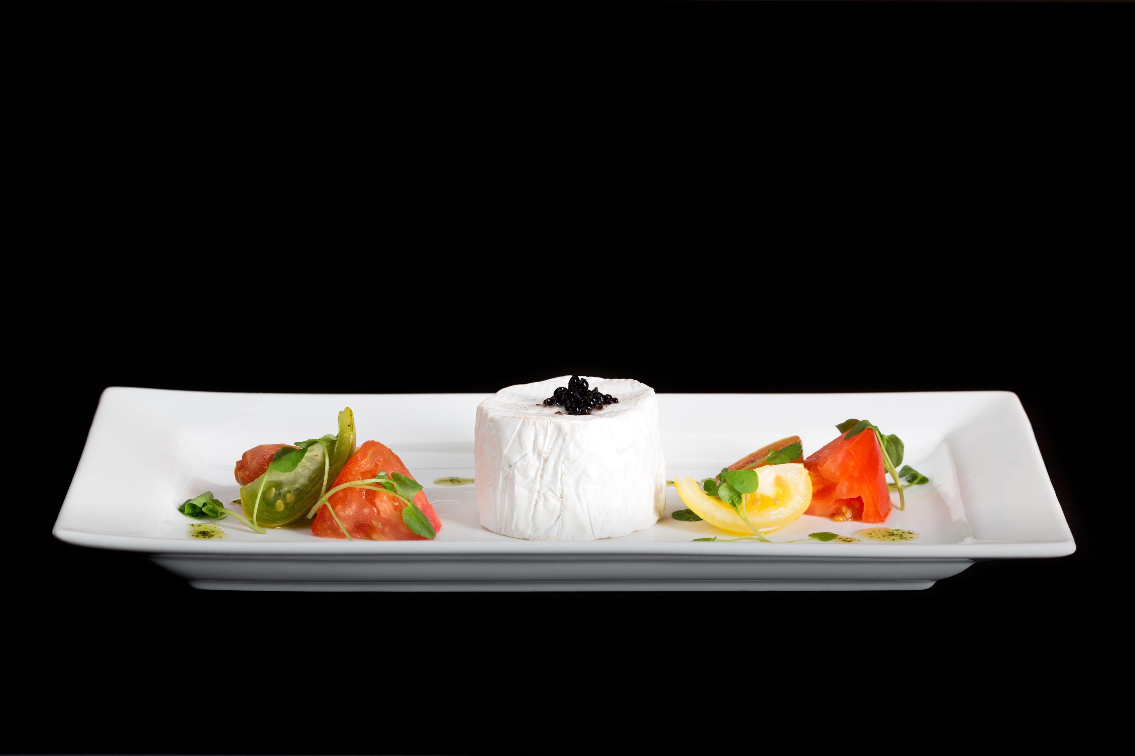 A white rectangular plate with a central round white cheese topped with black caviar, surrounded by various fresh vegetables.