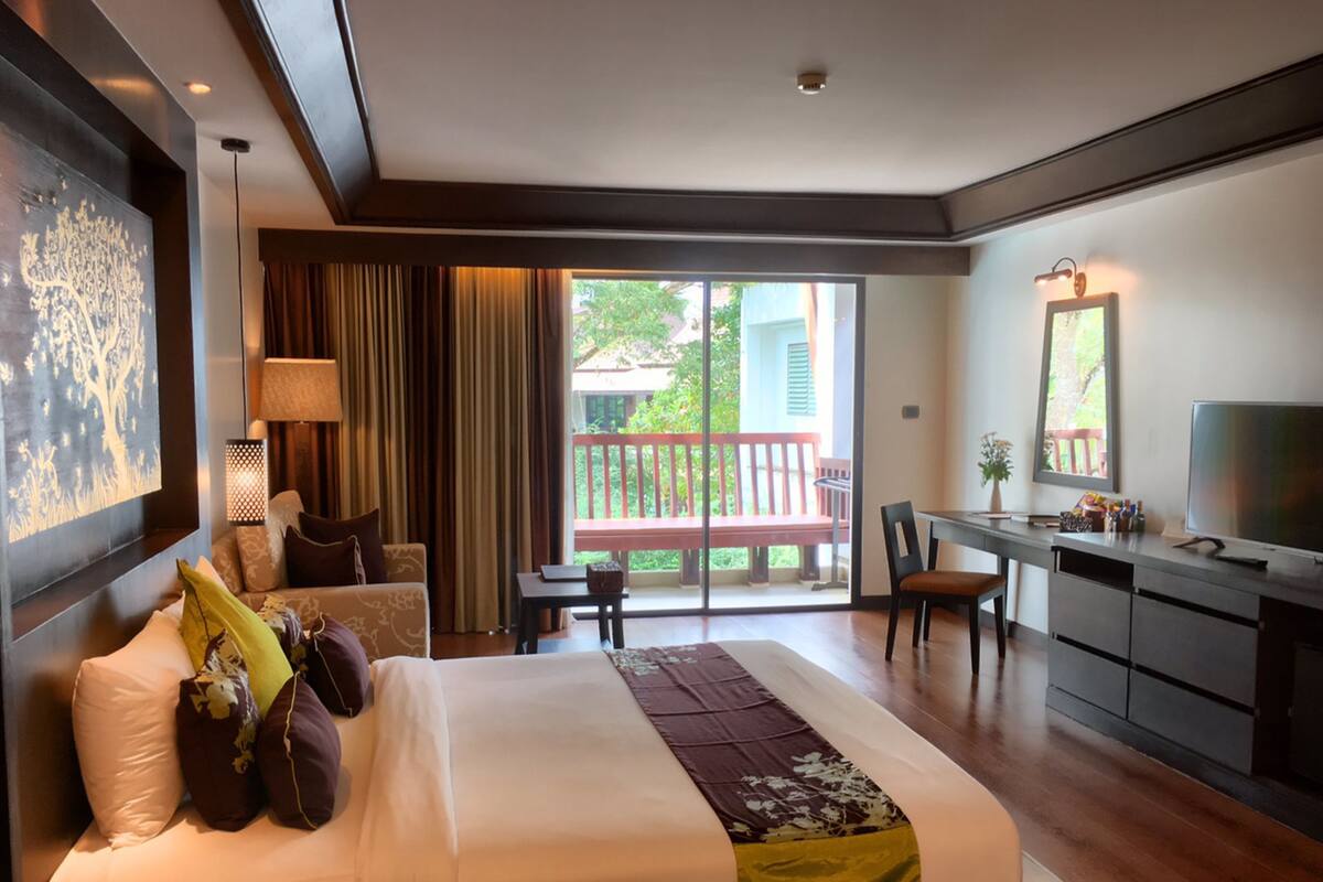 A hotel room with a bed, a desk, a chair, and a balcony with a view of greenery.