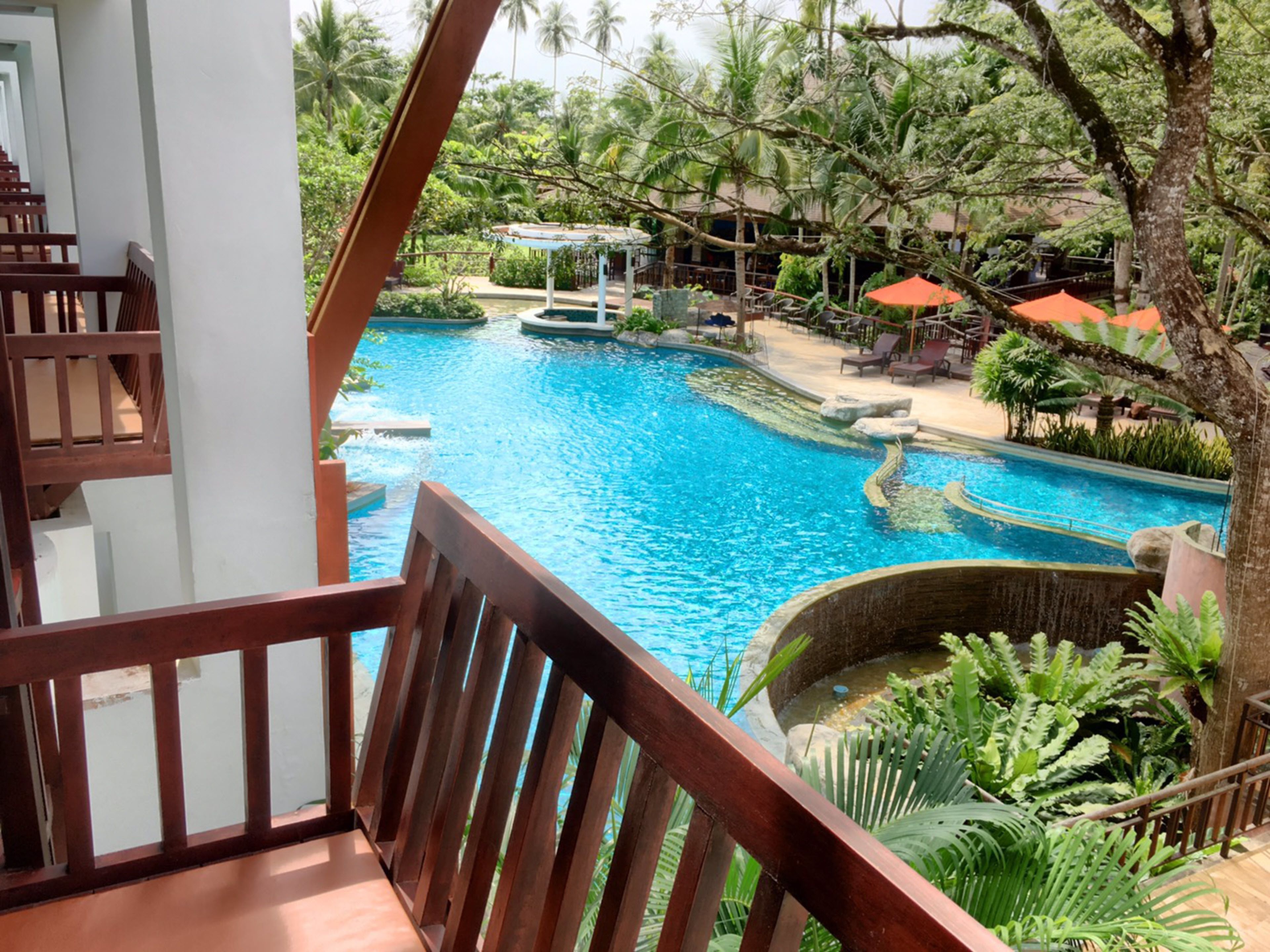 Vue depuis un balcon donnant sur une piscine, entourée de verdure et agrémentée d’un espace salon abrité.