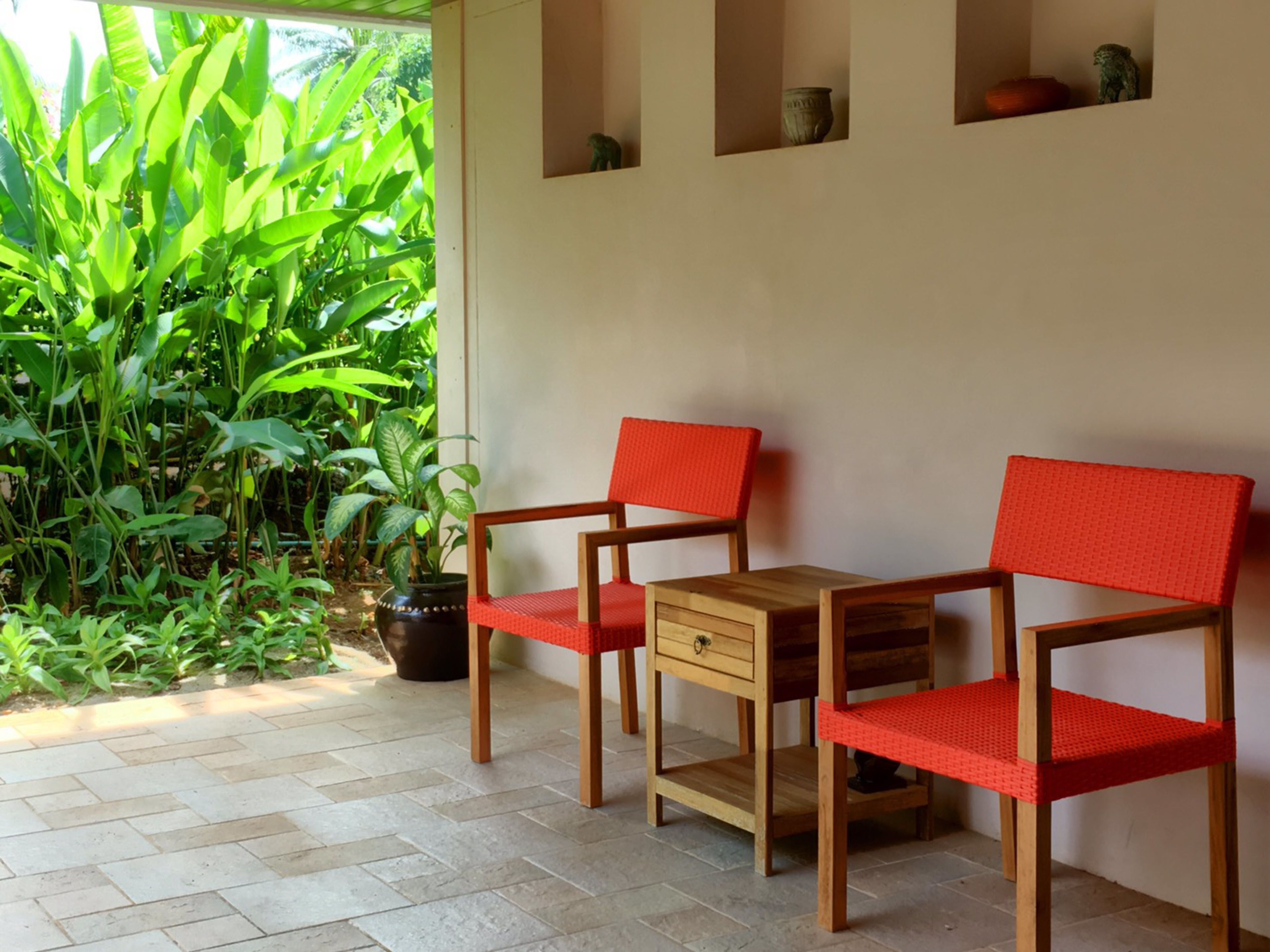 A patio with two wooden chairs and a small table, surrounded by lush green plants and a potted plant.