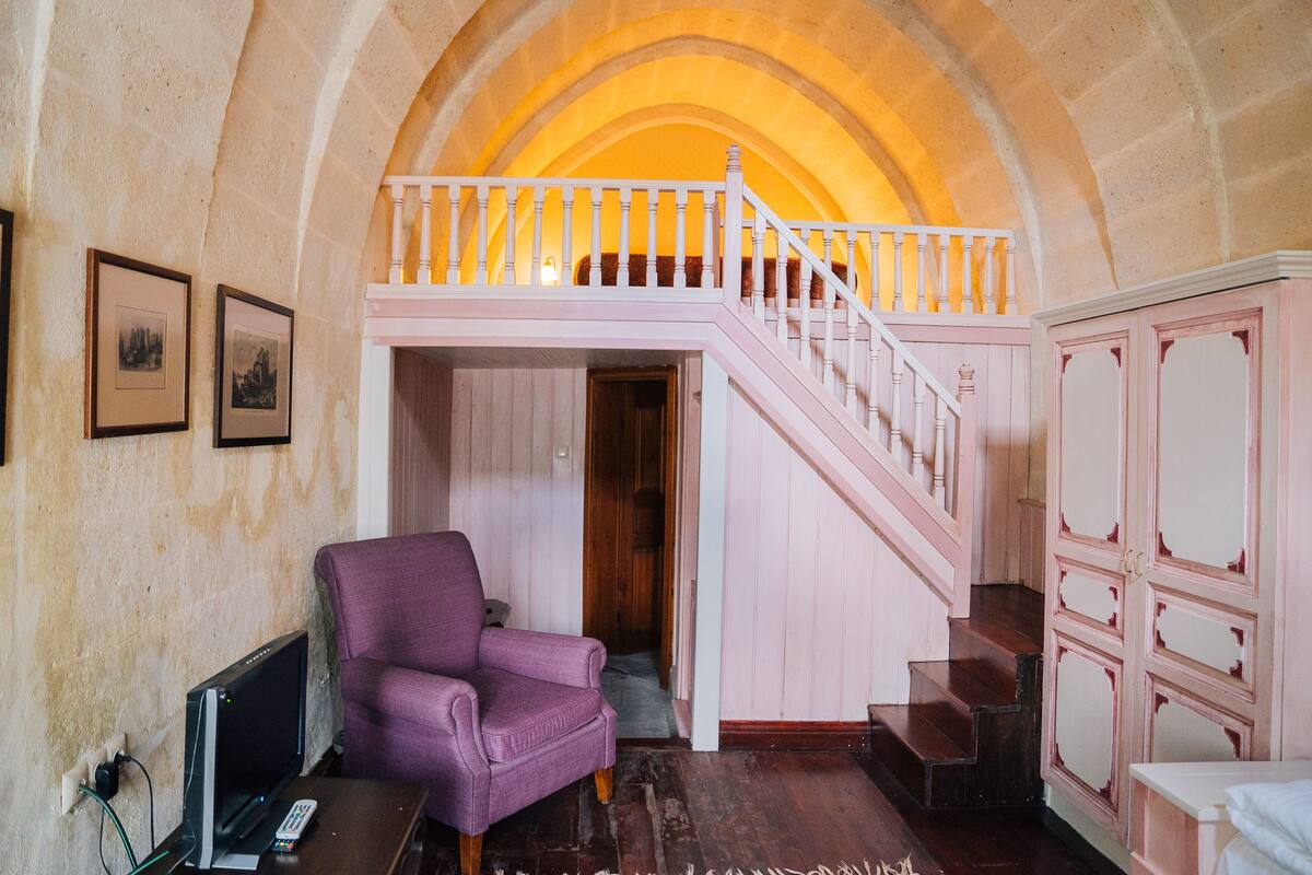 Ein Zimmer mit einer hohen gewölbten Decke, einem violetten Sessel, einem Fernseher und einer Treppe, die zu einer oberen Ebene führt.