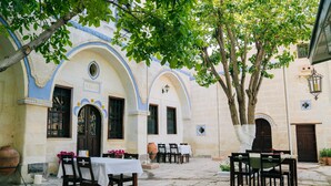An outdoor patio with tables and chairs, a large tree, and a building with blue and white arches.