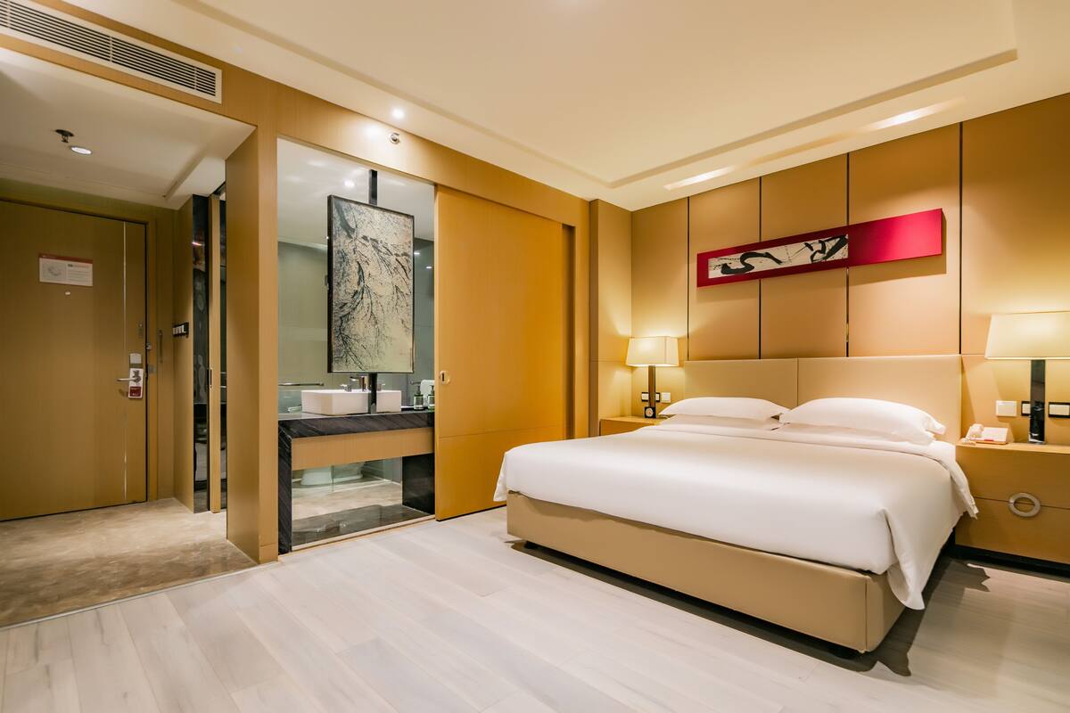 A modern hotel room with a large bed, a bathroom with a mirror and sink, and a red abstract painting on the wall.