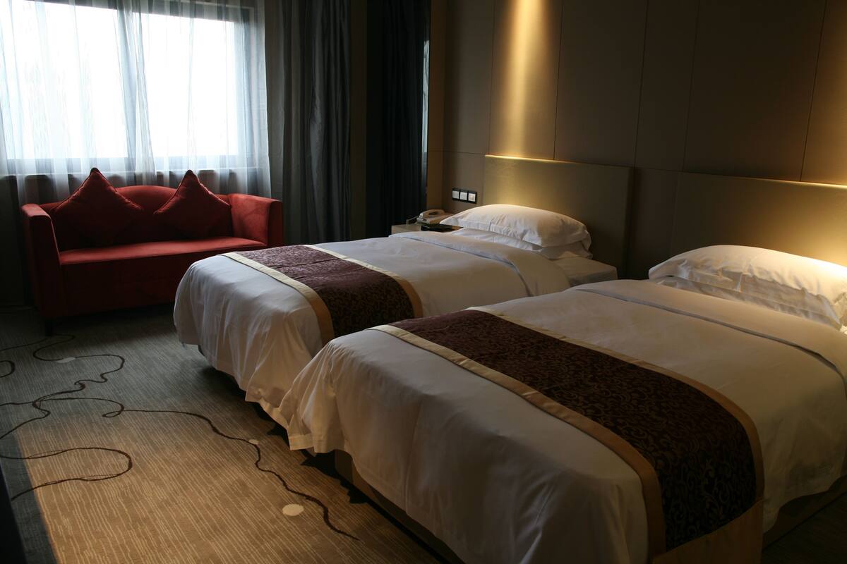 A hotel room with two beds, a red sofa, a bedside table with a phone, and a window with sheer curtains.