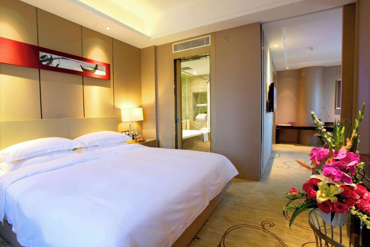 A hotel room with a large bed, a nightstand with a lamp, a mirror, and a floral arrangement.
