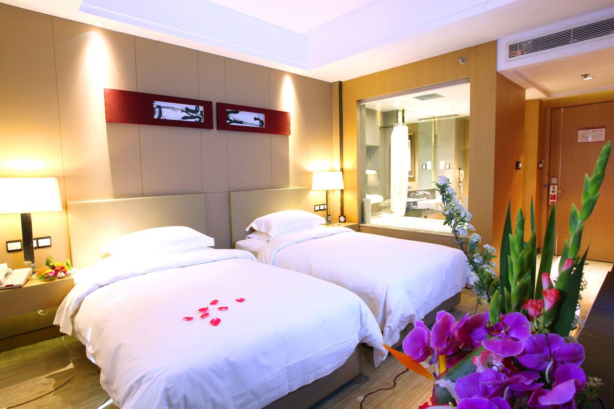 A hotel room with two beds, a nightstand, a lamp, a mirror, and a floral arrangement.