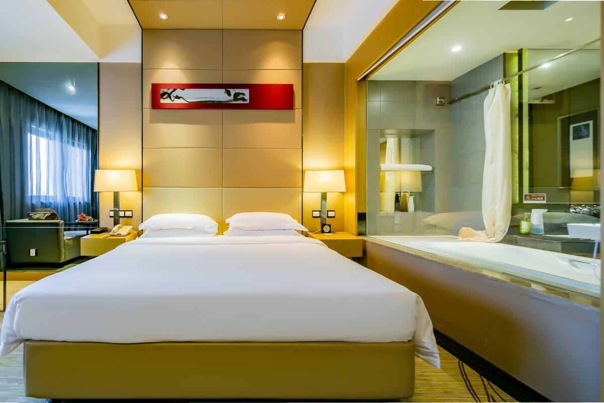A hotel room with a large bed, a sofa, a desk with a lamp, a nightstand, a bathroom with a glass shower, and a wall-mounted artwork.