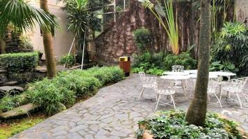 A stone-paved patio with white metal chairs and tables, surrounded by lush greenery and a stone wall.