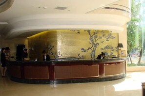A hotel reception area with a curved marble counter, a gold decorative wall, and a view of the outdoors. - Century Palace Hotel (Huizhou)