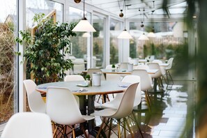 A conservatory with round tables and white chairs, large windows, and hanging pendant lights. - Hotel Stadt Gehrden (Gehrden)