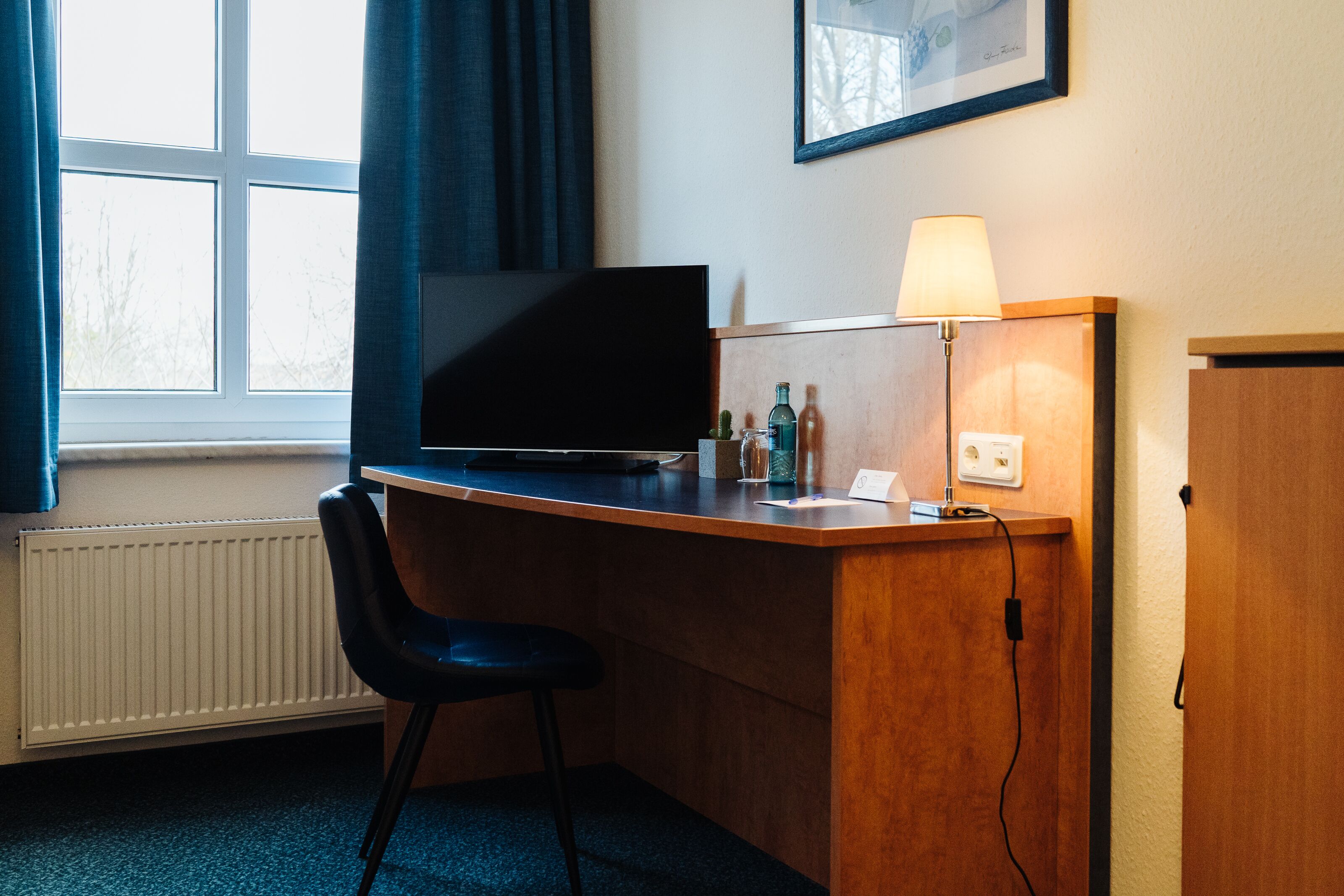 Habitación de hotel con un escritorio de madera, una silla negra, televisor, lámpara y ventana con cortinas.