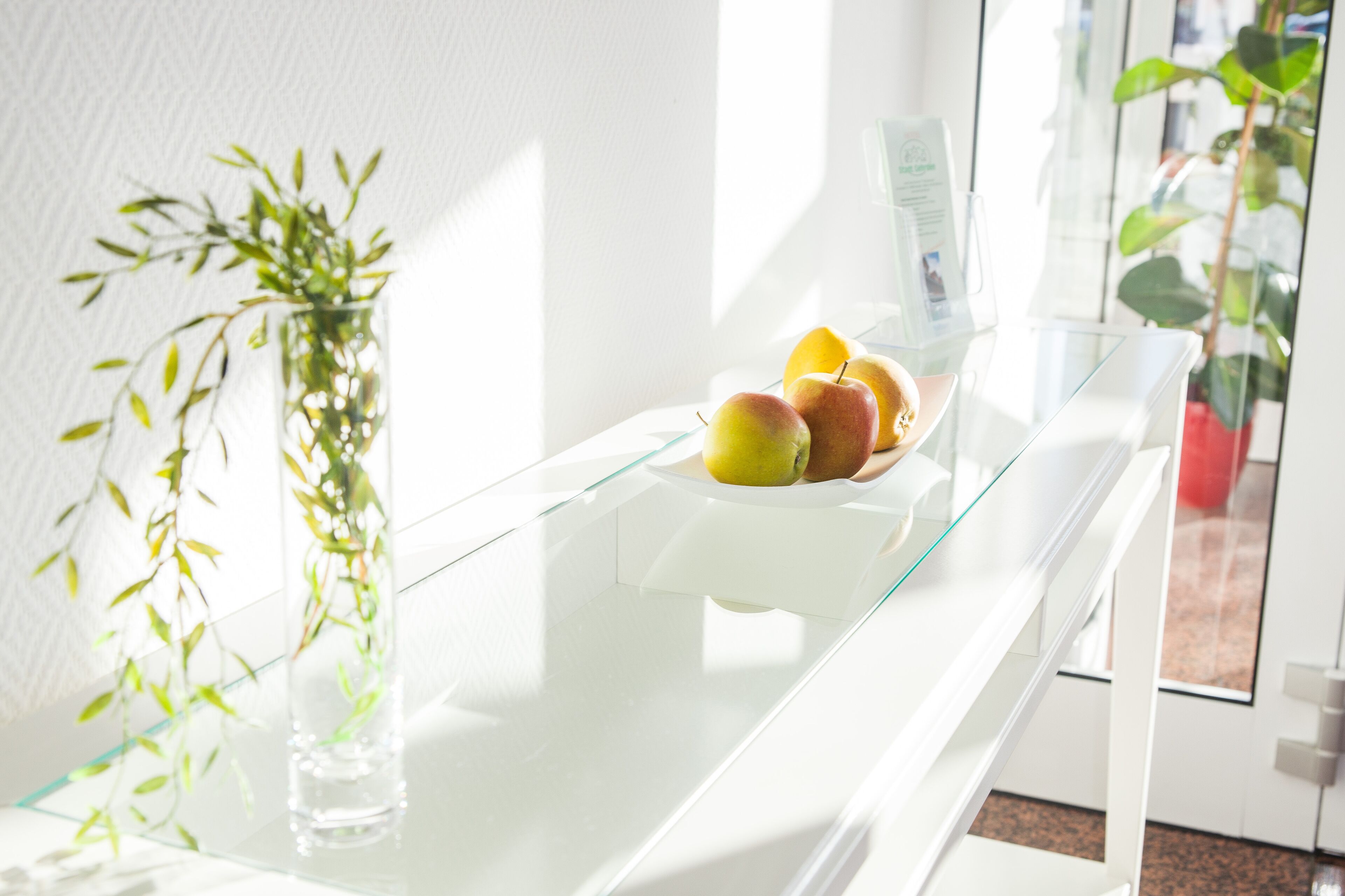 A glass-top table with a bowl of apples and a glass vase with green leaves.