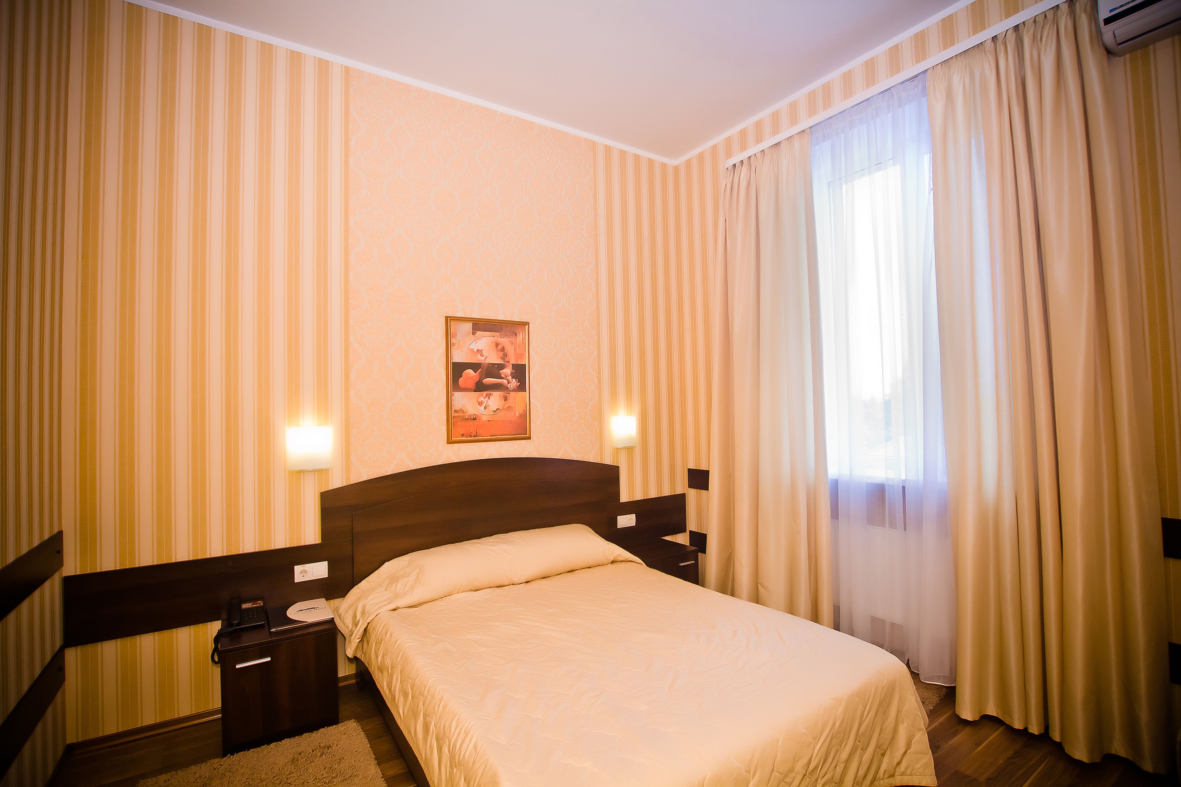 Habitación de hotel con cama, mesitas de noche, una mesita con lámpara, un cuadro en la pared y cortinas en las ventanas.