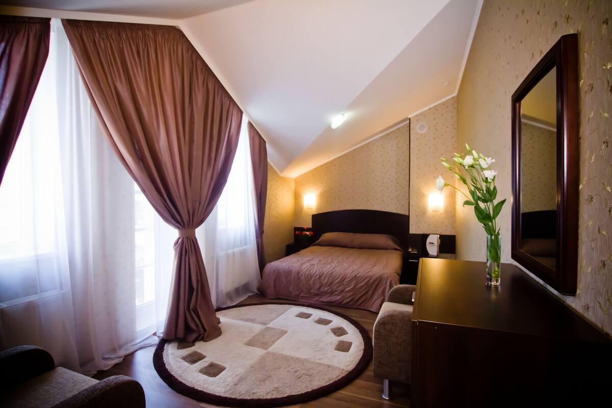 A bedroom with a bed, a round rug, a dresser, a mirror, and a nightstand with a lamp.