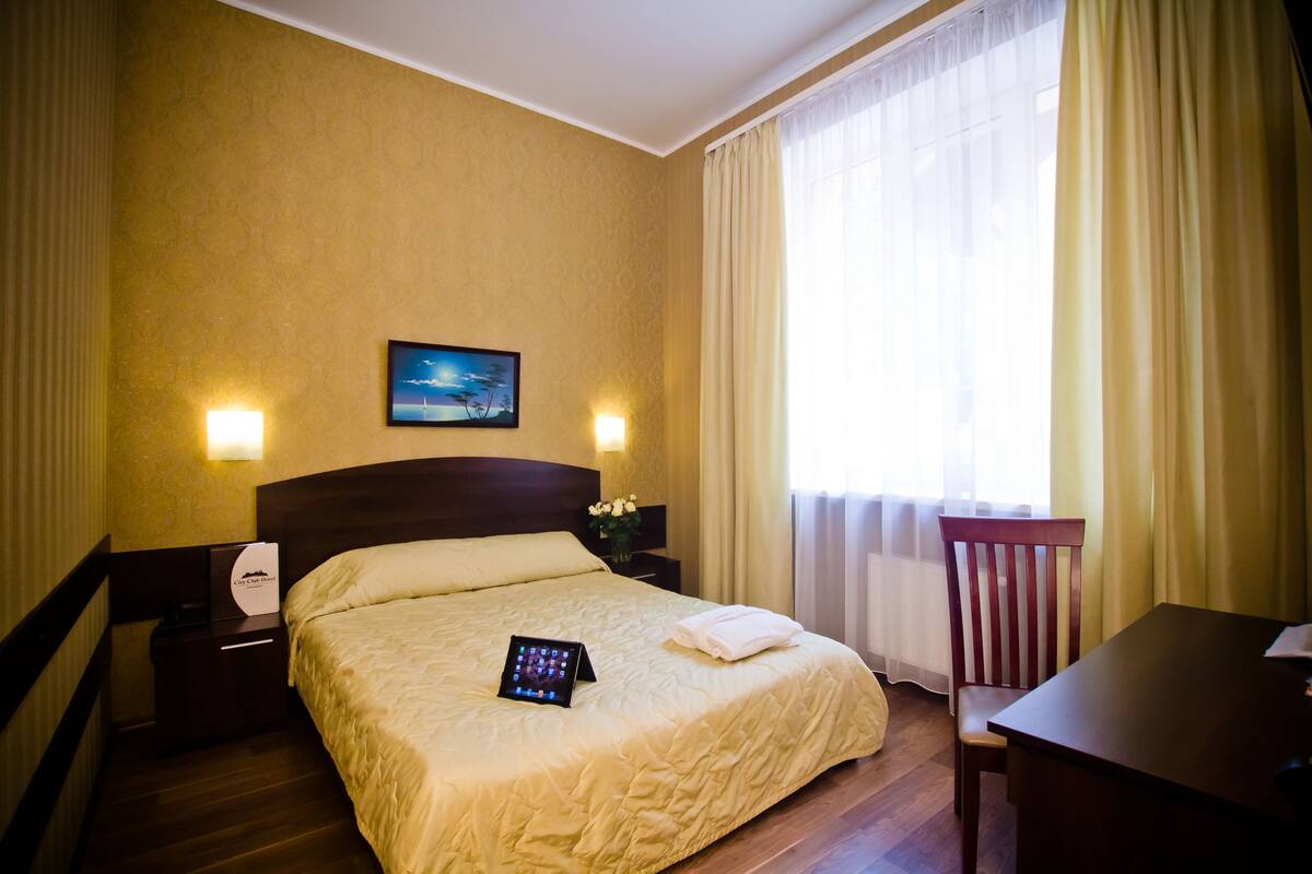 A hotel room with a bed, bedside table, lamp, and a view of the outdoors through a window.