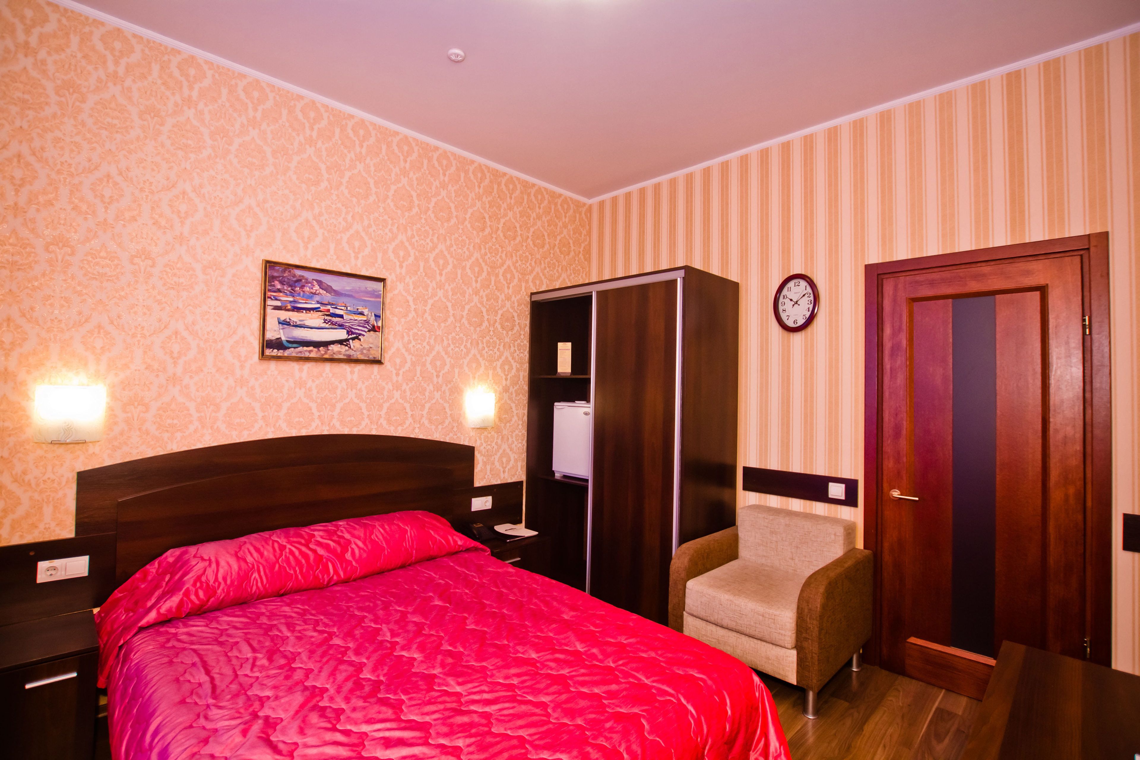A hotel room with a bed, a chair, a wardrobe, a clock, and a wall-mounted light.