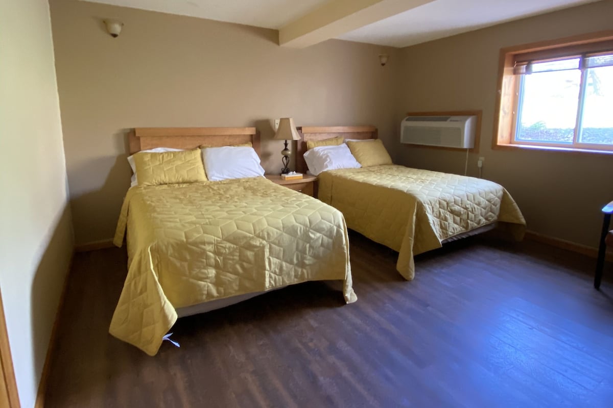Una habitación con dos camas, suelo de madera, una lámpara en el techo, una ventana y un equipo de aire acondicionado.