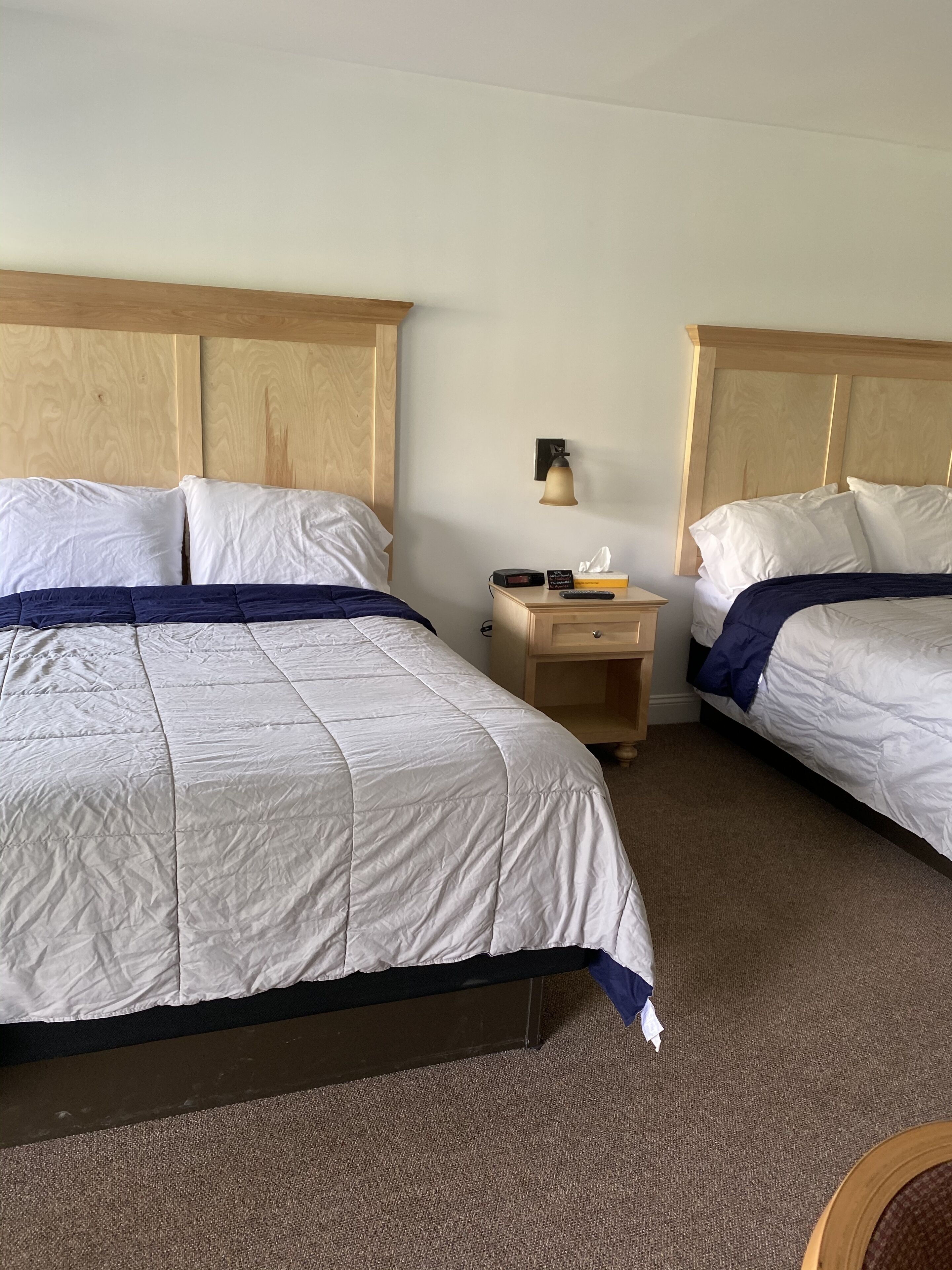 A hotel room with two beds, wooden headboards, a nightstand with a lamp, and a small wooden table.