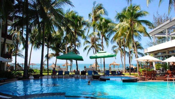 Una piscina con sillones de descanso y sombrillas, rodeada de palmeras y vista al océano.