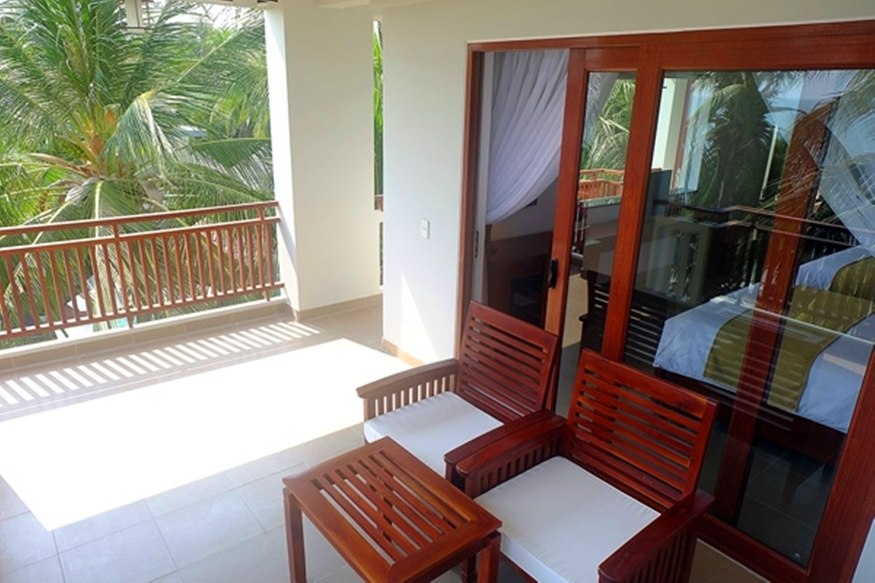 A balcony with wooden chairs and a table, a sliding glass door, and a view of palm trees.