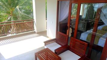 A balcony with wooden chairs and a table, a sliding glass door, and a view of palm trees.