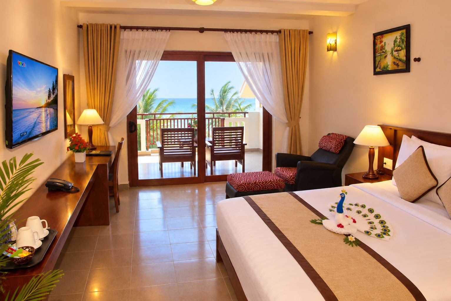 Habitación de hotel con cama, escritorio, sillón, sofá, balcón con vista al océano y un cuadro en la pared.