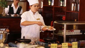 Un chef con uniforme blanco y un gorro alto prepara comida en una cocina donde se ven distintos utensilios y productos.