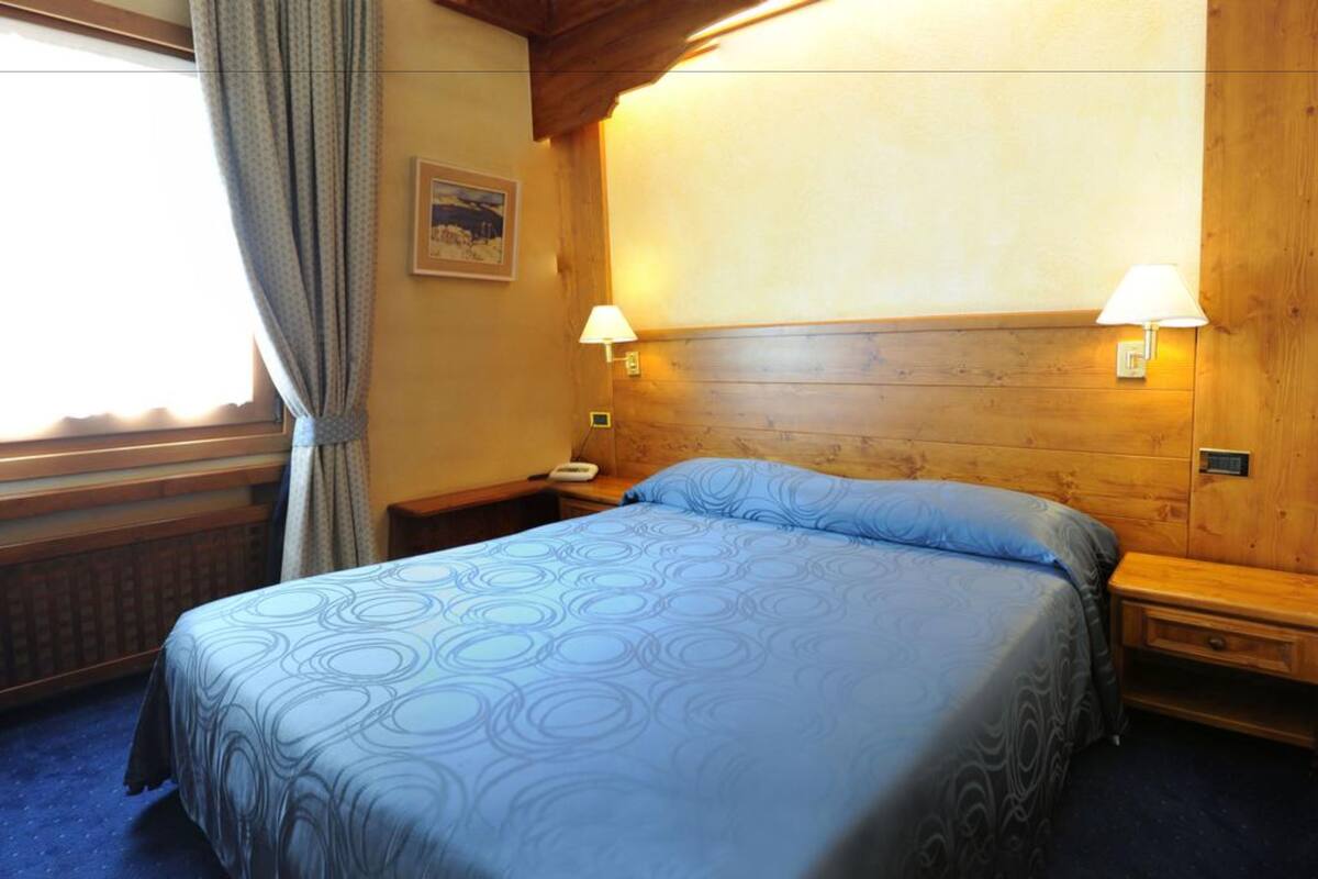 Une chambre d’hôtel avec un lit, deux tables de chevet, une commode, une lampe, un tableau au mur et une fenêtre avec des rideaux.