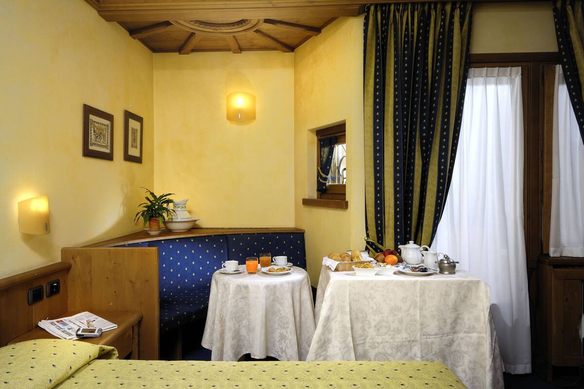 Une chambre d’hôtel avec une tête de lit en bois, un banc, deux petites tables rondes sur lesquelles sont disposés des éléments de petit-déjeuner, une lampe fixée au mur et une fenêtre avec des rideaux.