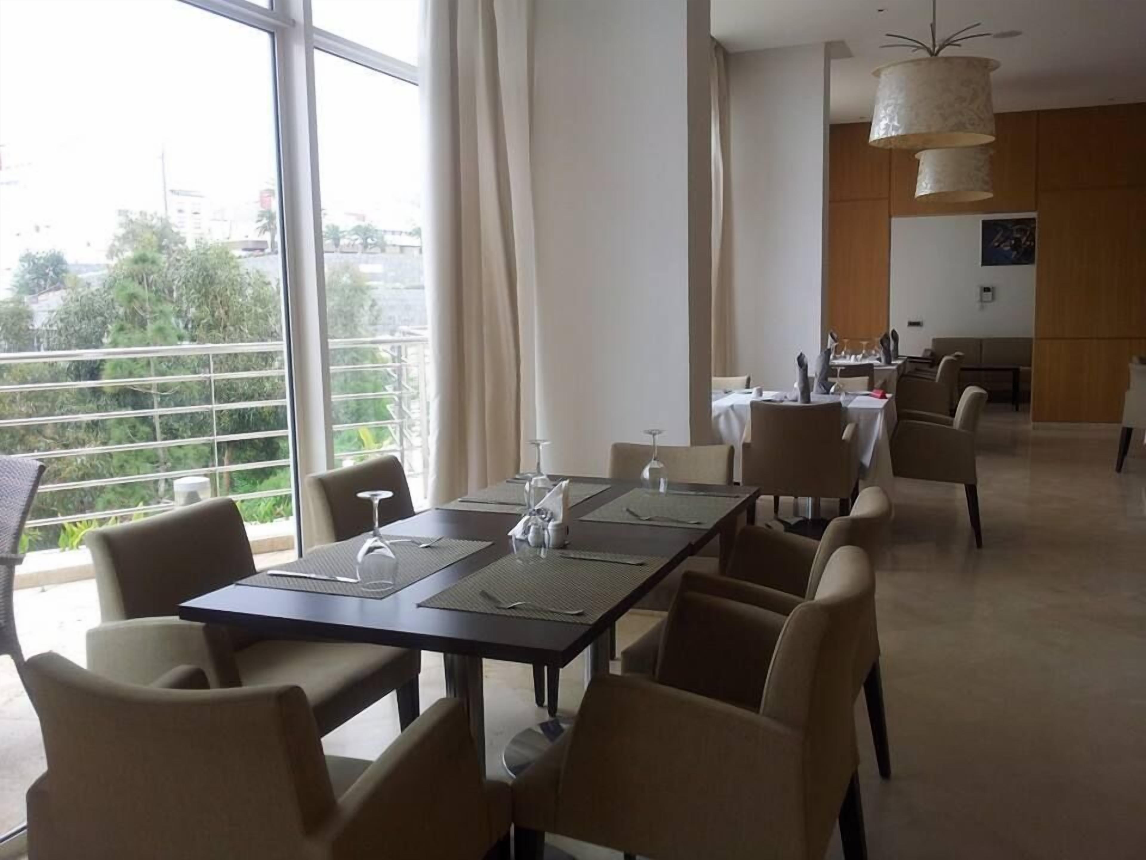 Een eetruimte met een lange tafel gedekt voor zes personen, omringd door stoelen, grote ramen met gordijnen en uitzicht op groen buiten.