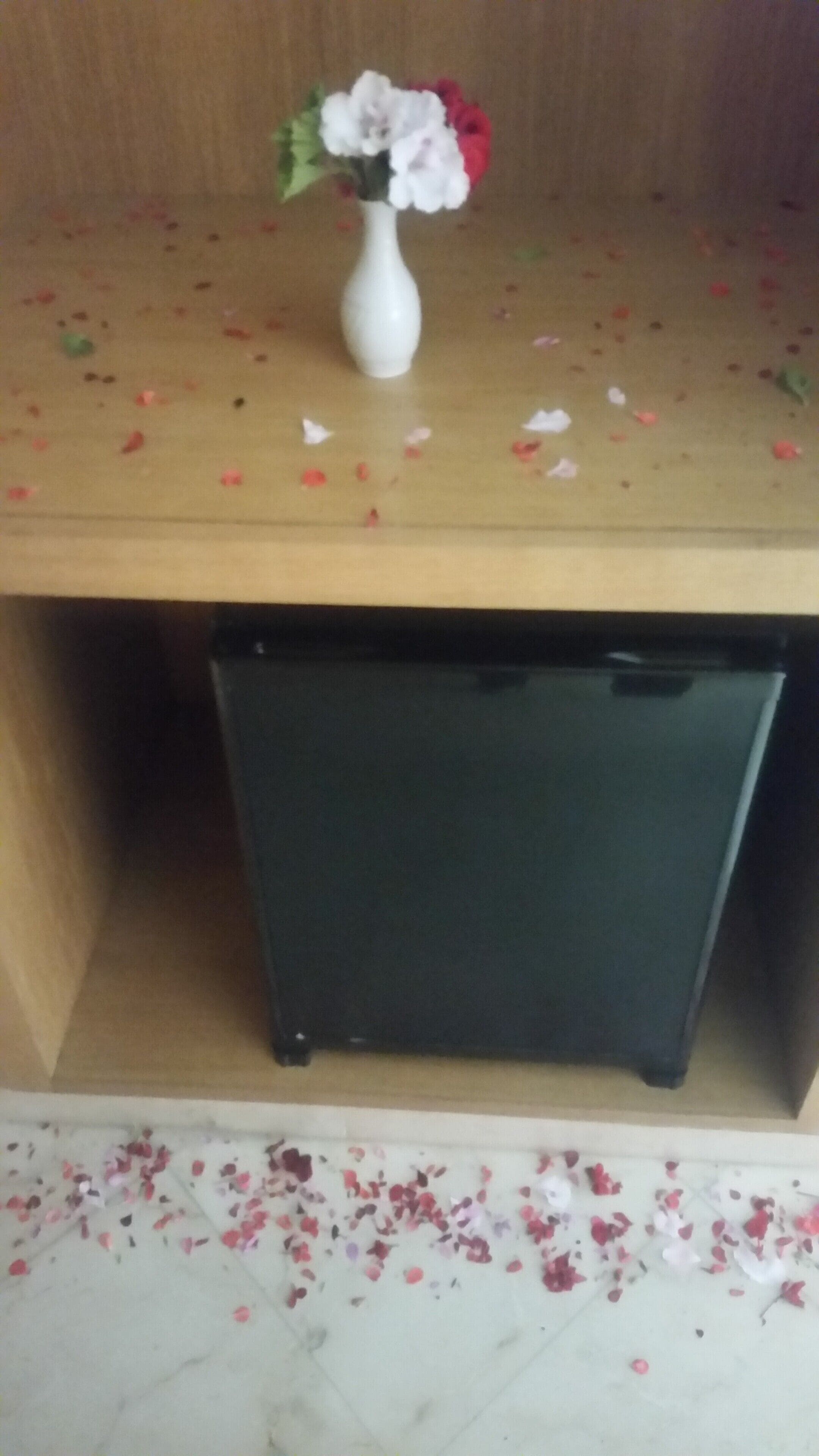 Una mesa de madera con un florero blanco que contiene flores, un mueble negro en la parte inferior y pétalos de flores esparcidos en el suelo.