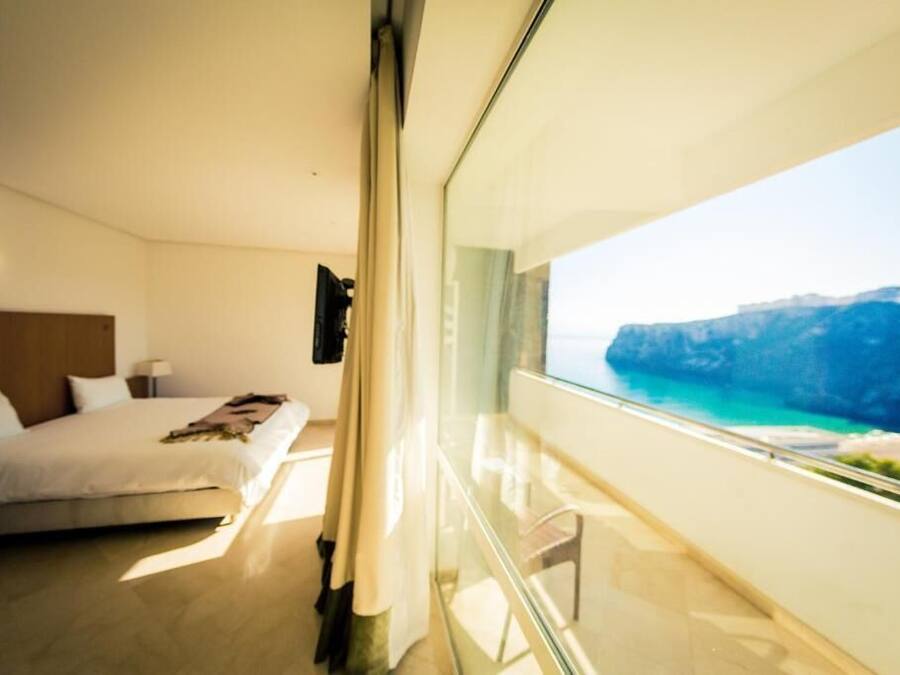 Een moderne slaapkamer met een groot bed, een nachtkastje en uitzicht op een kustlandschap door een glazen deur.